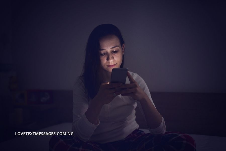Woman in pajamas sitting on a bed in dim lighting, illuminated by the glow of a handheld smartphone.