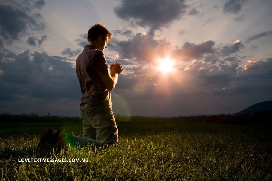 Man kneeling in tall grass with clasped hands facing the sun. Overcast sky. Distant hills.